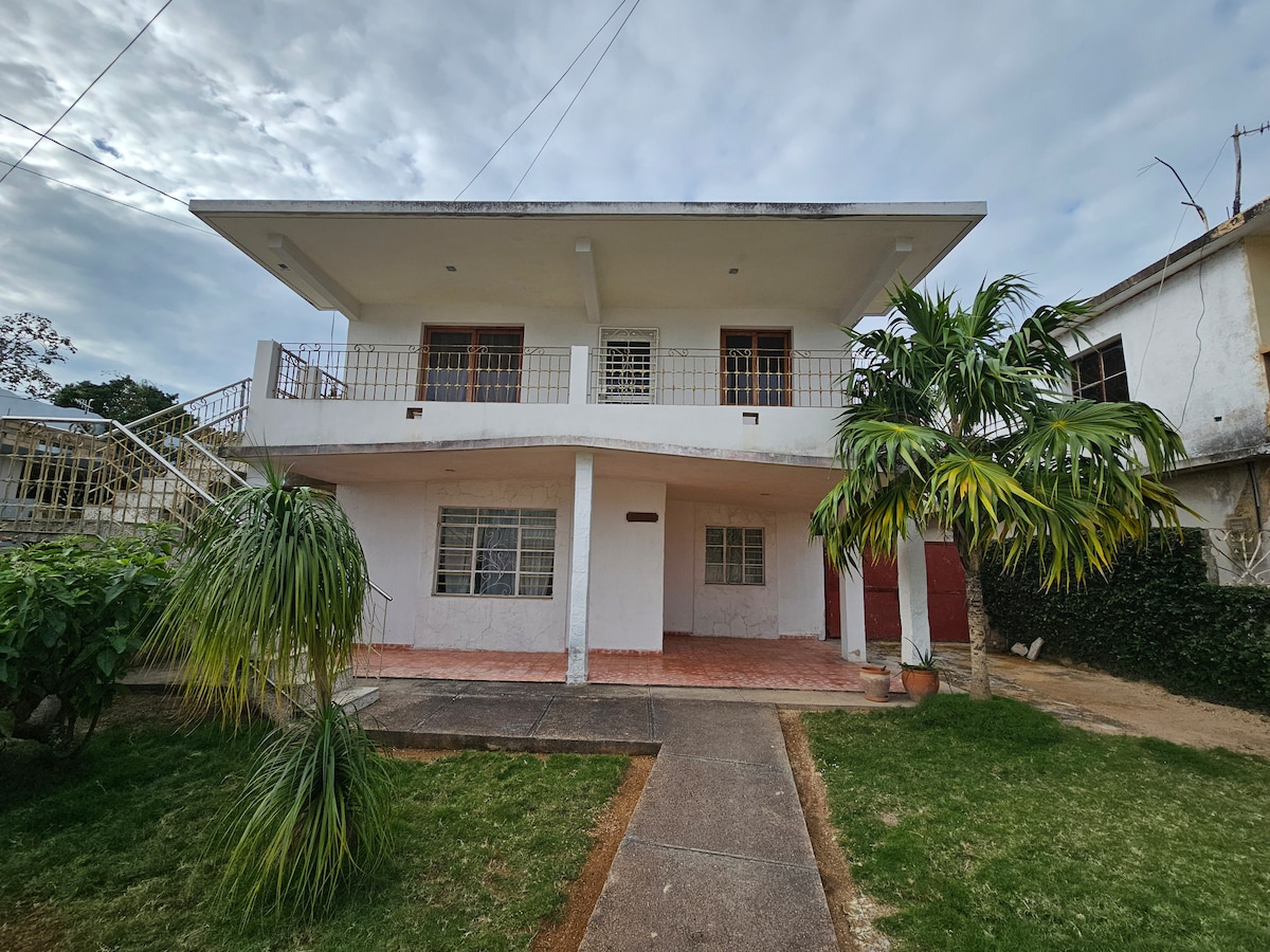 The exterior of La Casa Sueca is displayed, featuring a two-story building with a wide balcony and large windows. A spacious front patio is visible, surrounded by greenery. The entrance is framed by a couple of palm trees and a neatly maintained lawn.