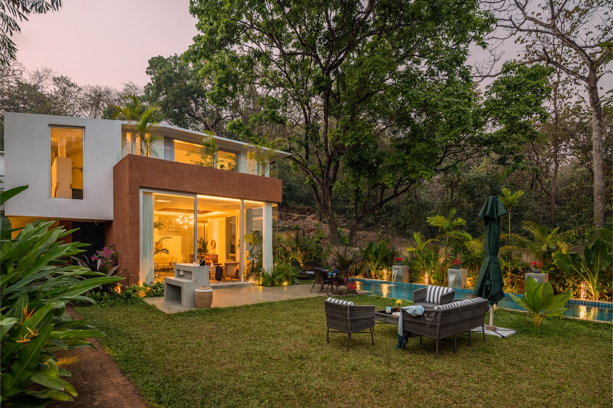 The modern villa is surrounded by lush greenery, featuring large glass windows that invite natural light. A serene pool reflects the surrounding plants, while outdoor seating arrangements offer a relaxing area for guests to enjoy the landscape.