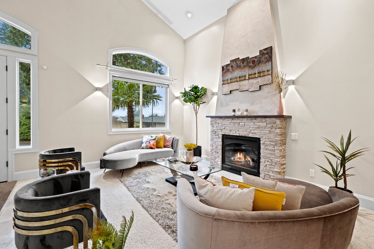 A stylish living area is enhanced by large windows allowing natural light to illuminate the space. A cozy seating arrangement includes a curved sofa and two armchairs, all oriented towards a modern fireplace with a stone facade. A coffee table sits in the center, complemented by decorative plants.