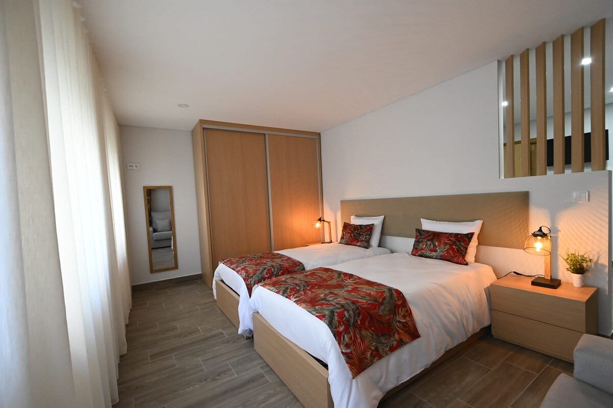 A bright bedroom is equipped with two spacious beds, dressed in linens featuring warm colors. A wooden wardrobe occupies one side, and a mirror stands nearby. Soft natural light filters through the sheer curtains, enhancing the overall ambiance of the room.
