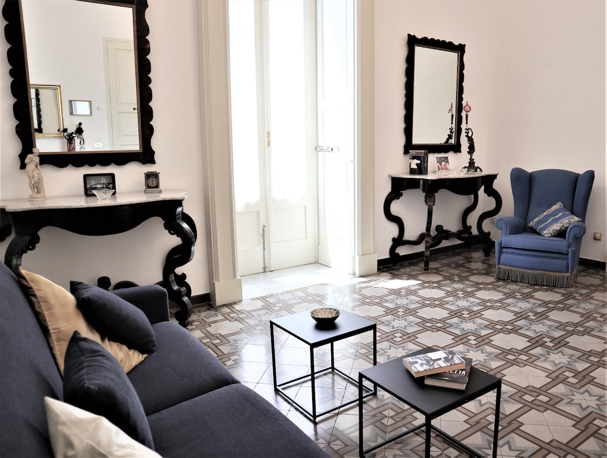 A spacious seating area is presented with a dark blue sofa and two small tables. An elegant blue armchair complements the furnishings. The room features large windows that allow natural light to fill the space, with decorative mirrors reflecting the room's charm.