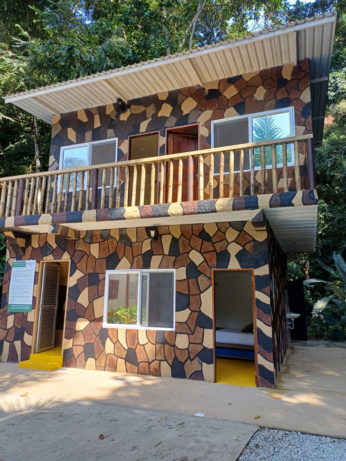 A two-story building is designed with an exterior of natural stone patterns, blending harmoniously with the surrounding greenery. The upper balcony features wooden railings, while large windows on both levels allow light to enter. A cozy sleeping area is visible through the open window.