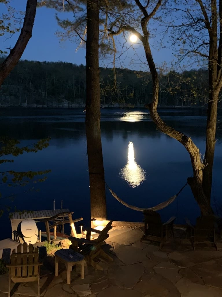 A serene lakeside scene is illuminated by moonlight, reflecting off the still water. Wooden chairs surround a stone patio, and a hammock is seen nearby, inviting relaxation. Tall trees frame the view, enhancing the peaceful ambiance of the outdoor space.