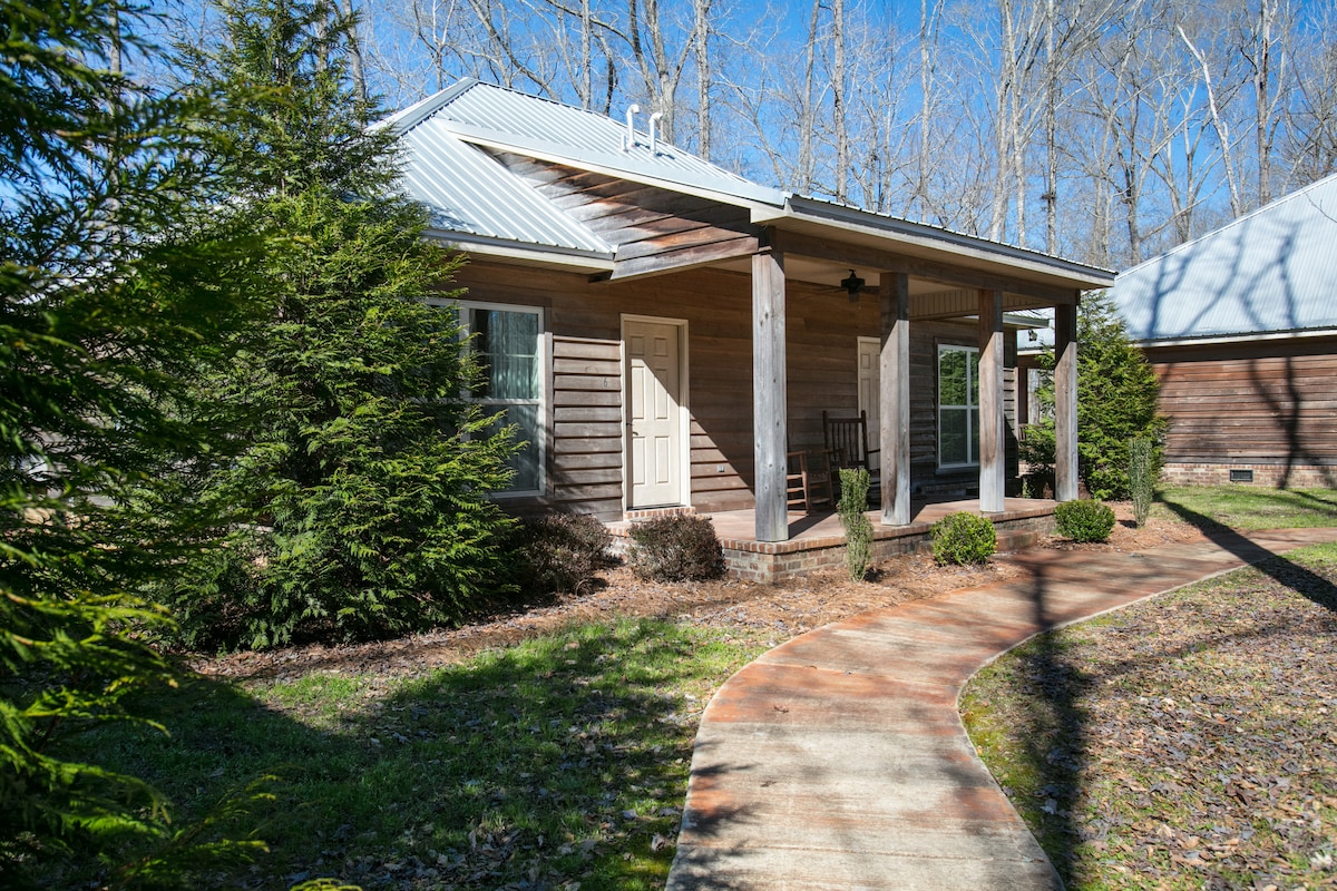 A cabin's exterior is showcased, emphasizing a welcoming entrance with a light-colored door and a rocking chair on the porch. Surrounded by well-maintained greenery, a winding pathway invites guests to explore. Sunlight filters through the trees, illuminating the peaceful setting.