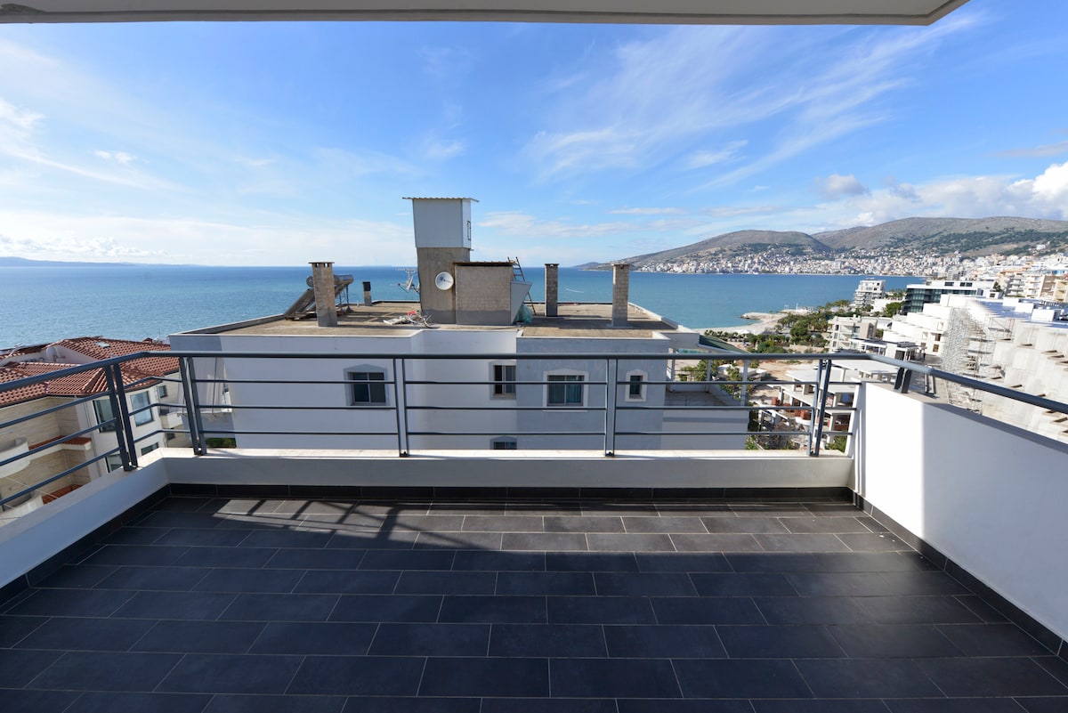 A spacious balcony offers sweeping views of the Ionian Sea, with the coastline and distant hills visible. The outdoor area features a sleek tiled floor and a modern railing, creating an inviting space for relaxation and enjoyment of the scenic surroundings.