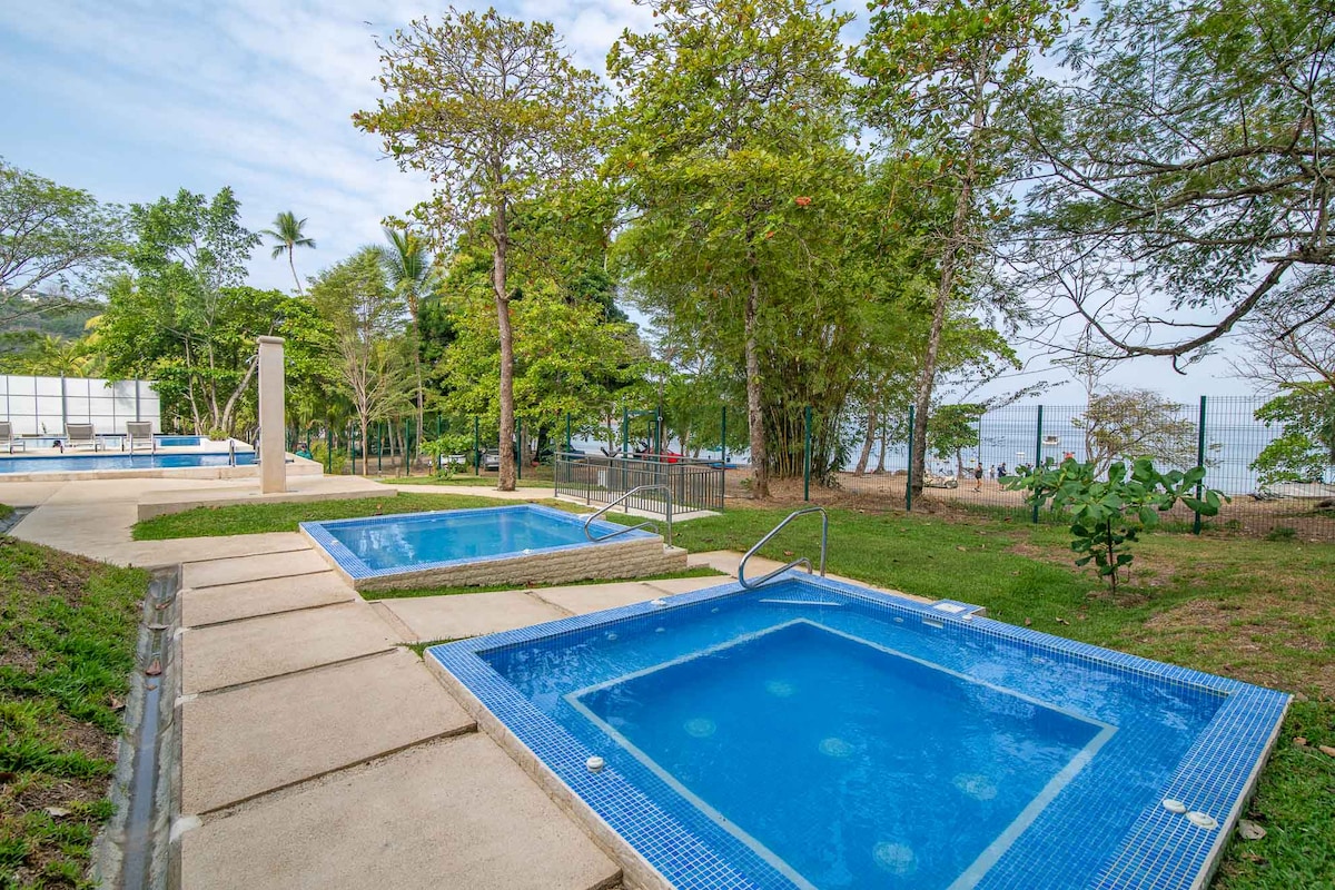 Two inviting jacuzzis are set in a serene outdoor space surrounded by lush greenery, with neatly paved pathways leading to each unit. The tranquil atmosphere is enhanced by the nearby ocean visible in the distance, inviting relaxation in a peaceful environment.