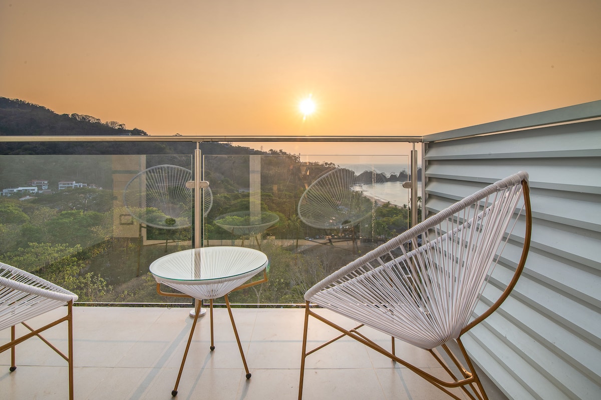 A serene balcony offers a view of the sun setting over the horizon. Two stylish chairs are positioned around a small, round table, creating an inviting space for relaxation. The glass railing enhances the openness of the area, allowing for unobstructed scenic views.