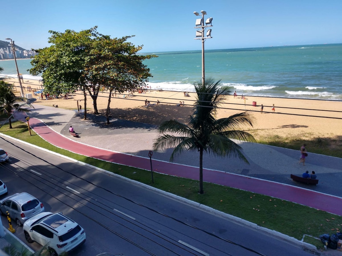 A view from the apartment showcases a sandy beach and a promenade lined with palm trees. The ocean extends into the distance, with gentle waves lapping at the shore. People can be seen enjoying the beach, while a pathway for walking and cycling runs parallel to the shore.