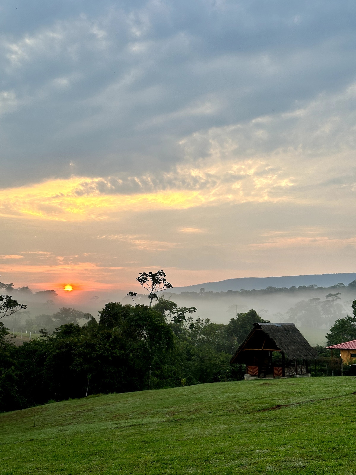 A serene landscape captures a sunrise, illuminating the sky with soft orange and pink hues. A rustic cabin is positioned among gentle hills and misty trees, with lush green grass in the foreground, creating a tranquil environment.