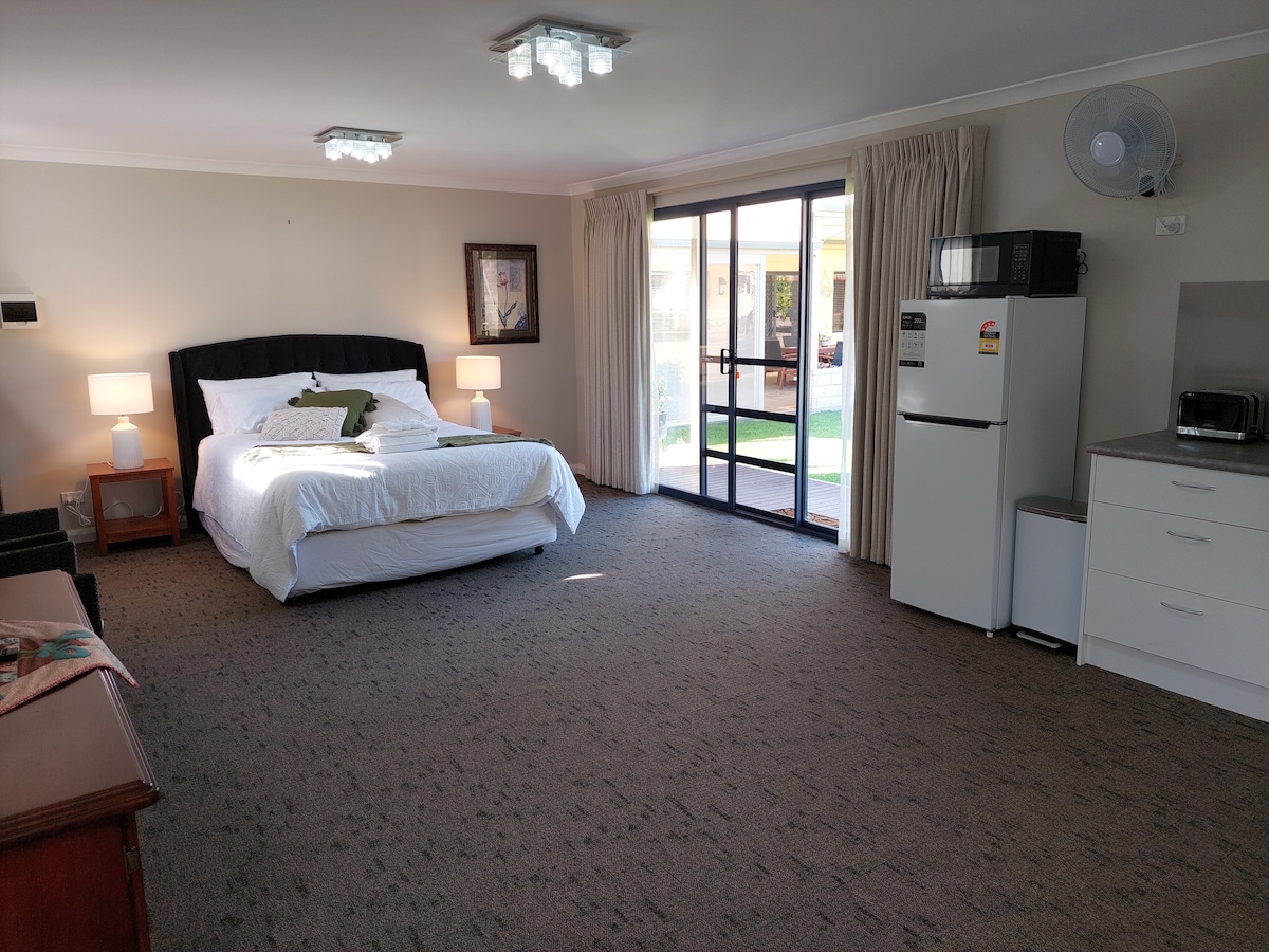A spacious room is featured, showcasing a large bed with white linens and decorative pillows. Natural light filters through glass sliding doors, which lead to an outdoor area. A small kitchenette is equipped with a refrigerator and microwave, complemented by nightstands and lamps.