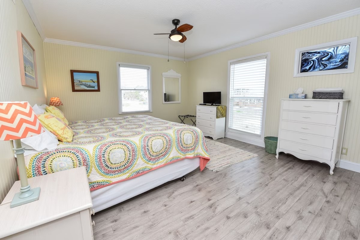 A spacious bedroom features a comfortable bed with a colorful patterned blanket. Natural light enters through two windows adorned with blinds. A dresser and a mirror enhance the room's layout, while a small desk is positioned near a window, offering a functional workspace.