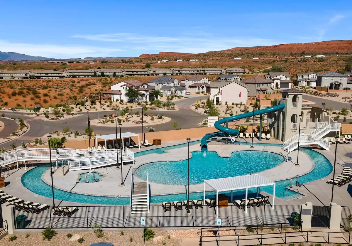 The image showcases a large community pool with a winding slide and a lazy river feature. Sun loungers are arranged around the pool area, complemented by scenic views of surrounding hills and homes. Clear blue skies enhance the inviting outdoor space.