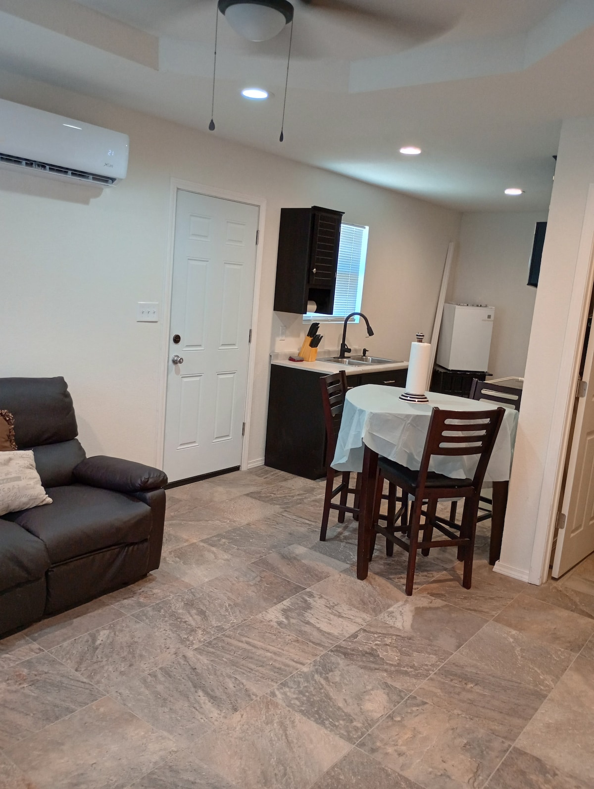 A compact living space features a dark leather sofa and a small table with four wooden chairs. A kitchenette is visible in the background, equipped with modern appliances. Natural light enters through a window, and a ceiling fan adds to the comfort of the room.