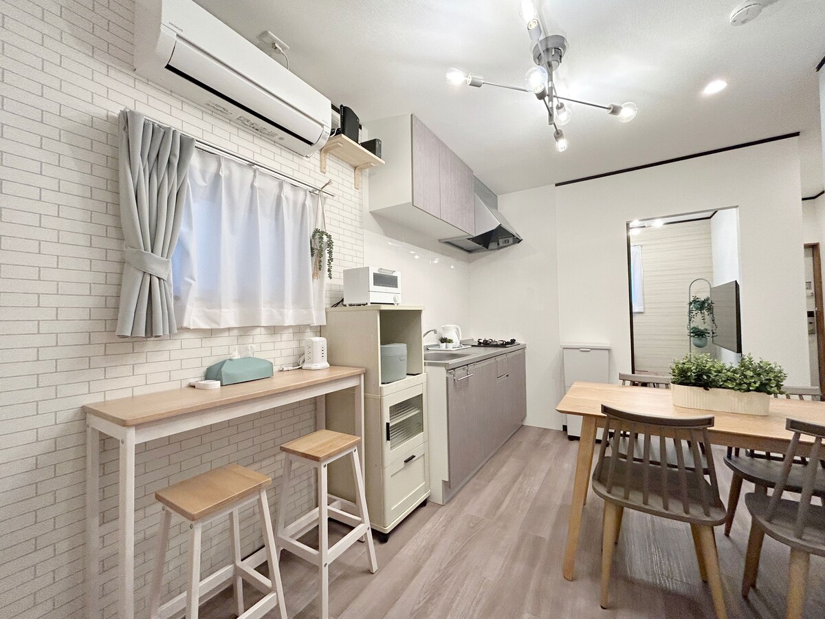 A bright kitchen area is highlighted by light-colored cabinetry and natural wood accents. A small dining table with four chairs is positioned next to an air conditioning unit. Kitchen appliances including a gas stove, microwave, and refrigerator are neatly arranged, creating a functional cooking space.