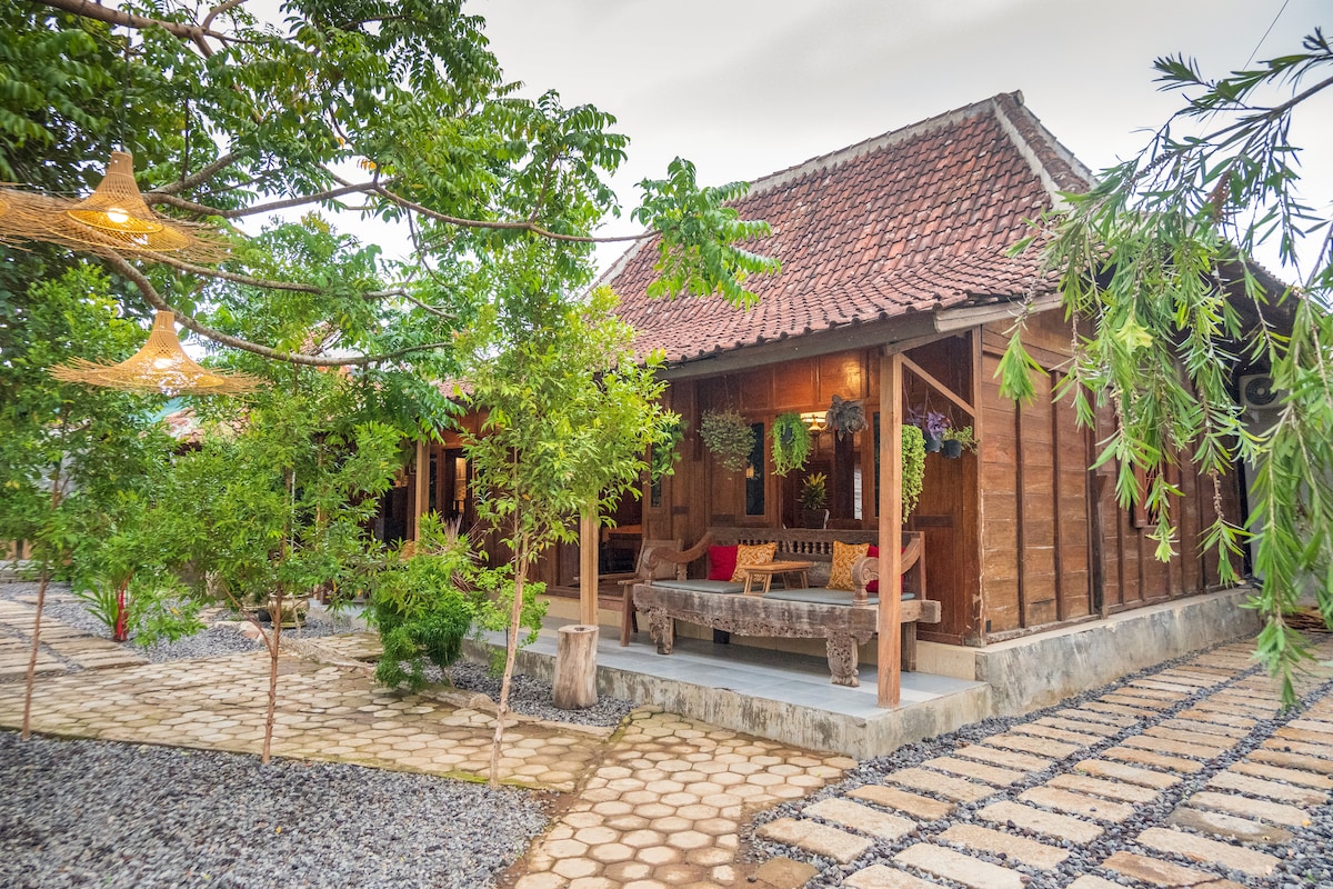 A traditional wooden structure with a thatched roof is surrounded by lush greenery. A stone pathway leads to a spacious porch decorated with outdoor seating and potted plants, creating a serene and inviting entrance.