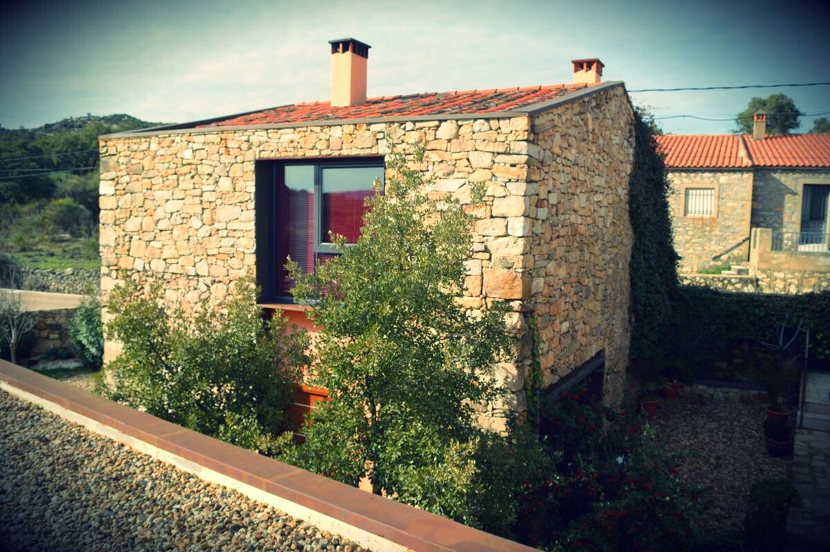 A rustic stone house is prominently featured, with large windows allowing natural light to illuminate the interior. Surrounding greenery adds a touch of nature, while red-tiled roofs of nearby buildings can be seen in the background, contributing to the rural ambiance.