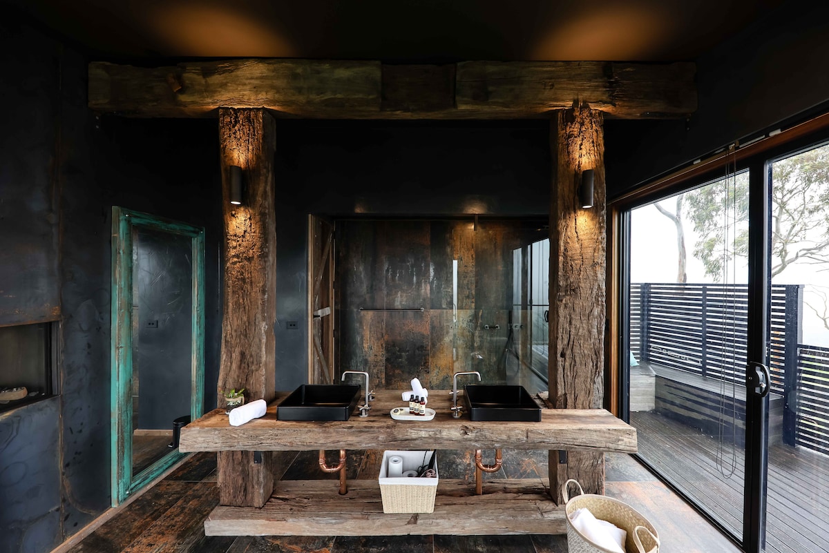A spacious bathroom features a rustic wooden double sink vanity supported by reclaimed wharf beams. The dark walls create a dramatic backdrop, while large glass panels allow natural light to fill the space, showcasing the outdoor views. Elegant lighting and decorative elements enhance the serene atmosphere.