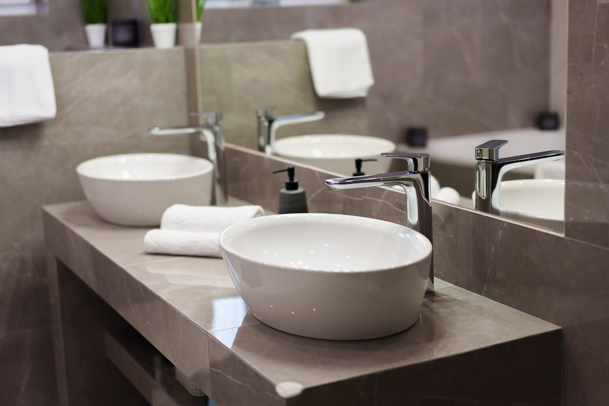A modern bathroom features dual sleek, oval sinks situated on a smooth gray countertop. Decorative plants are placed above the sinks, and white towels are neatly arranged nearby. Reflections in the mirror enhance the spacious feel of the area.