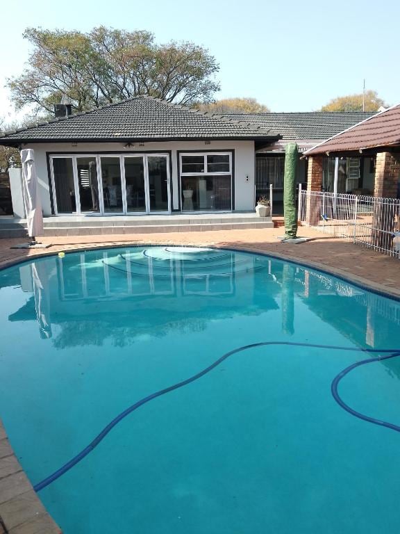 An outdoor swimming pool features calm blue water surrounded by a spacious patio area. Glass doors lead into the guest house, allowing for views of the interior. Lush greenery is visible in the background, contributing to a relaxed atmosphere.
