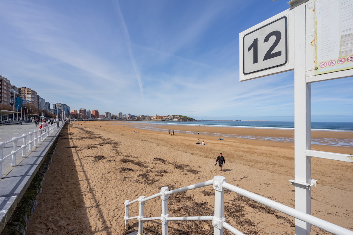 A sandy beach is viewed from a promenade, with a white sign indicating the number 12. Gentle waves caress the shoreline, while a few people are seen walking and relaxing along the beach. Buildings line the background, contrasting with the blue sky.