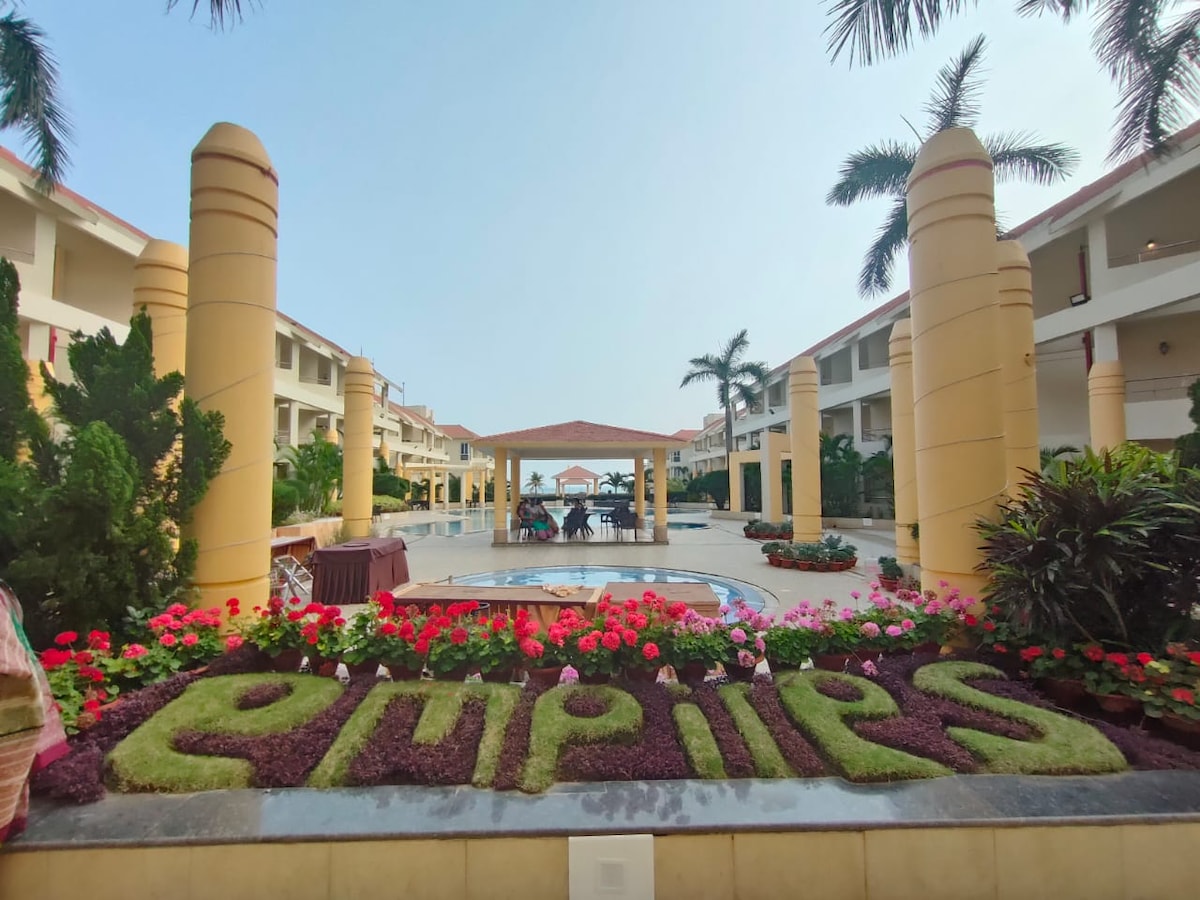 The entrance of Empires Hotel is showcased, featuring lush greenery and vibrant flower beds that spell 'Empires.' Towering yellow columns stand on either side, creating a welcoming pathway. The central courtyard includes a covered seating area, with hints of the beachfront visible in the background.