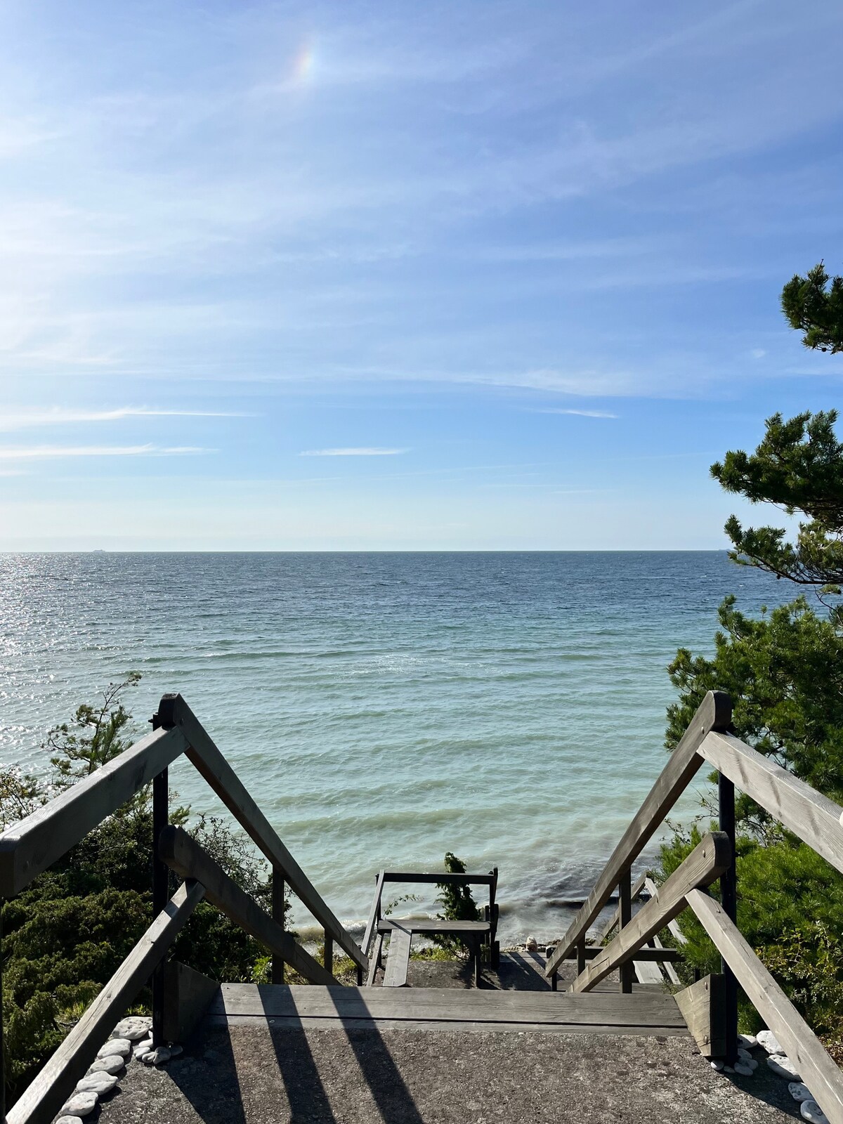 A wooden staircase leads down towards a calm sea, with gentle waves lapping at the shore. The clear sky above features soft clouds, and lush green foliage frames the scene on either side. The water displays varying shades of blue and green, inviting relaxation.