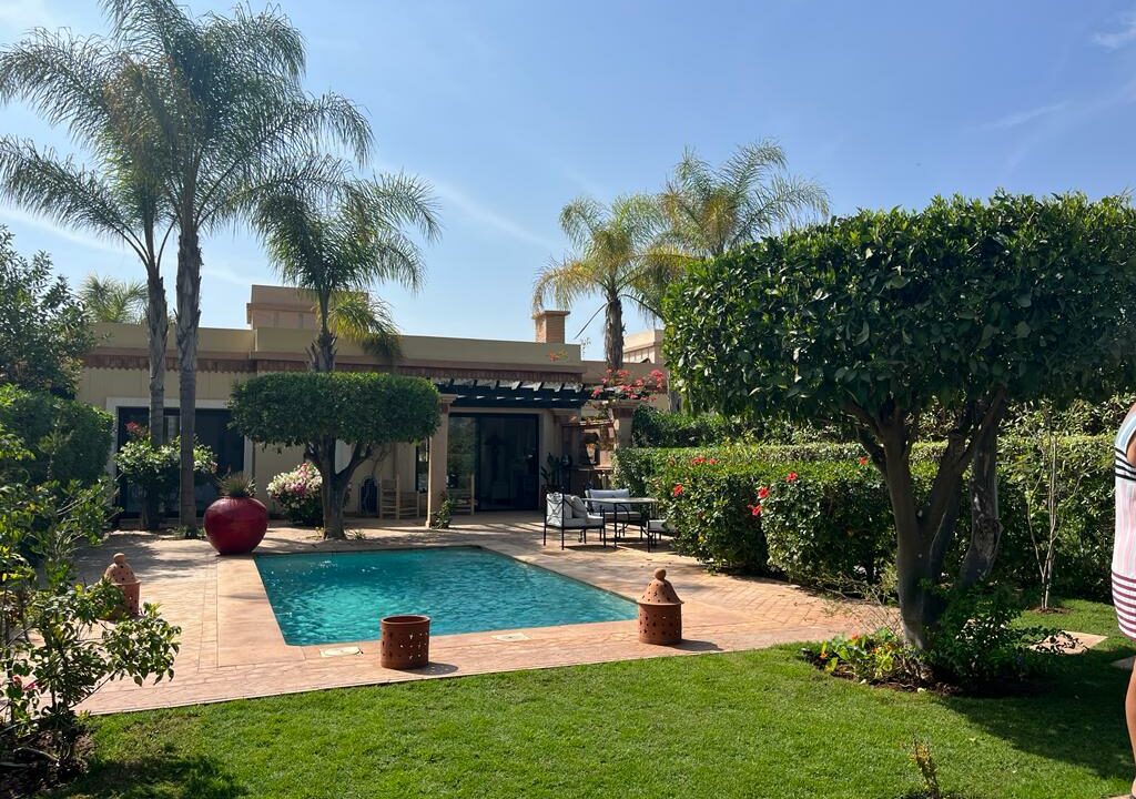 The outdoor area features a private swimming pool surrounded by lush greenery and neatly trimmed hedges. Comfortable seating arrangements are present near the pool, complemented by decorative planters. Palm trees provide shade under a clear blue sky, enhancing the natural ambiance of the villa.
