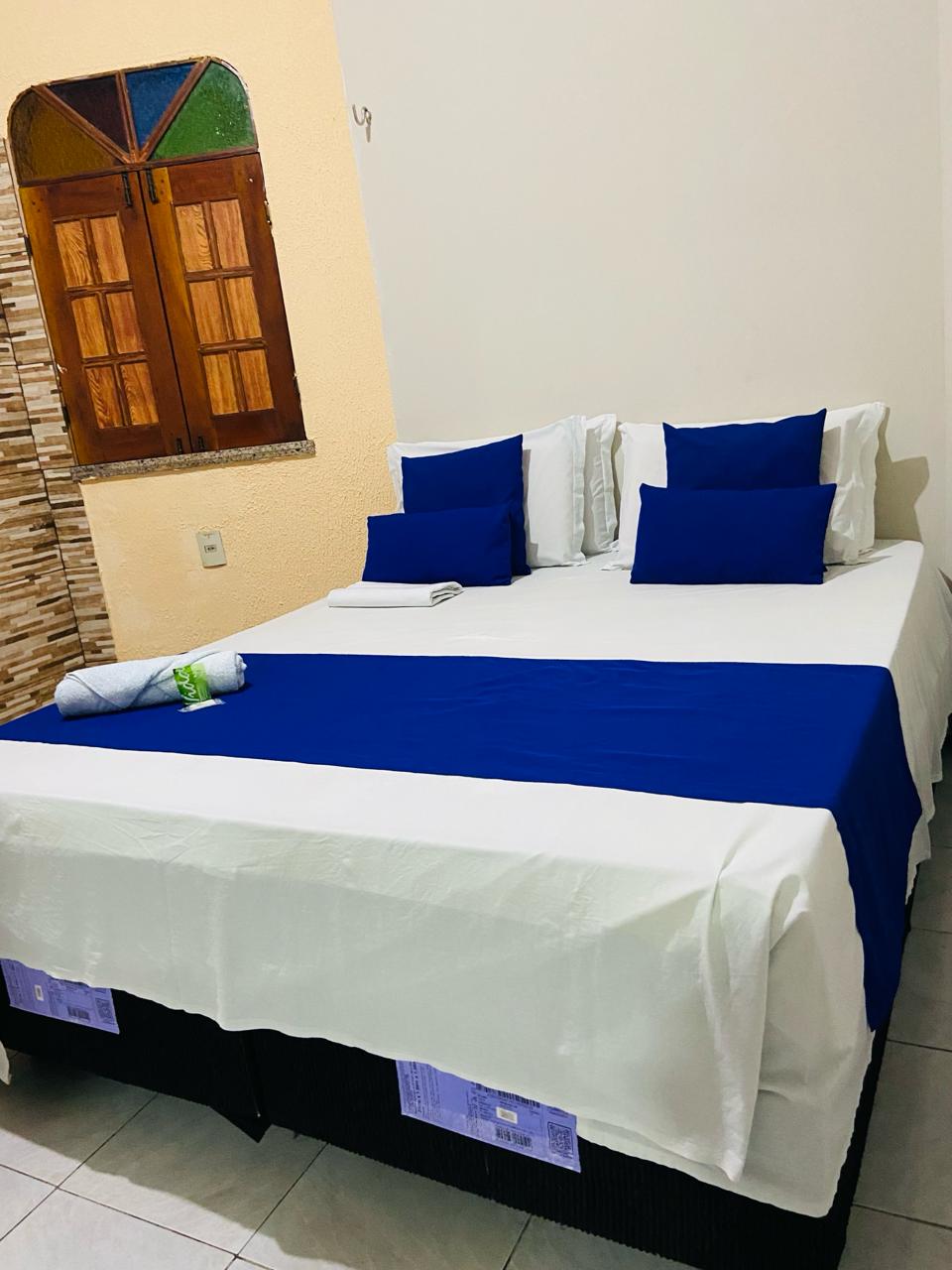 A spacious bed is adorned with white linens and decorative blue pillows. A neatly rolled towel and additional linens are placed at the foot of the bed. A window with wooden shutters provides natural light, enhancing the room's airy feel.