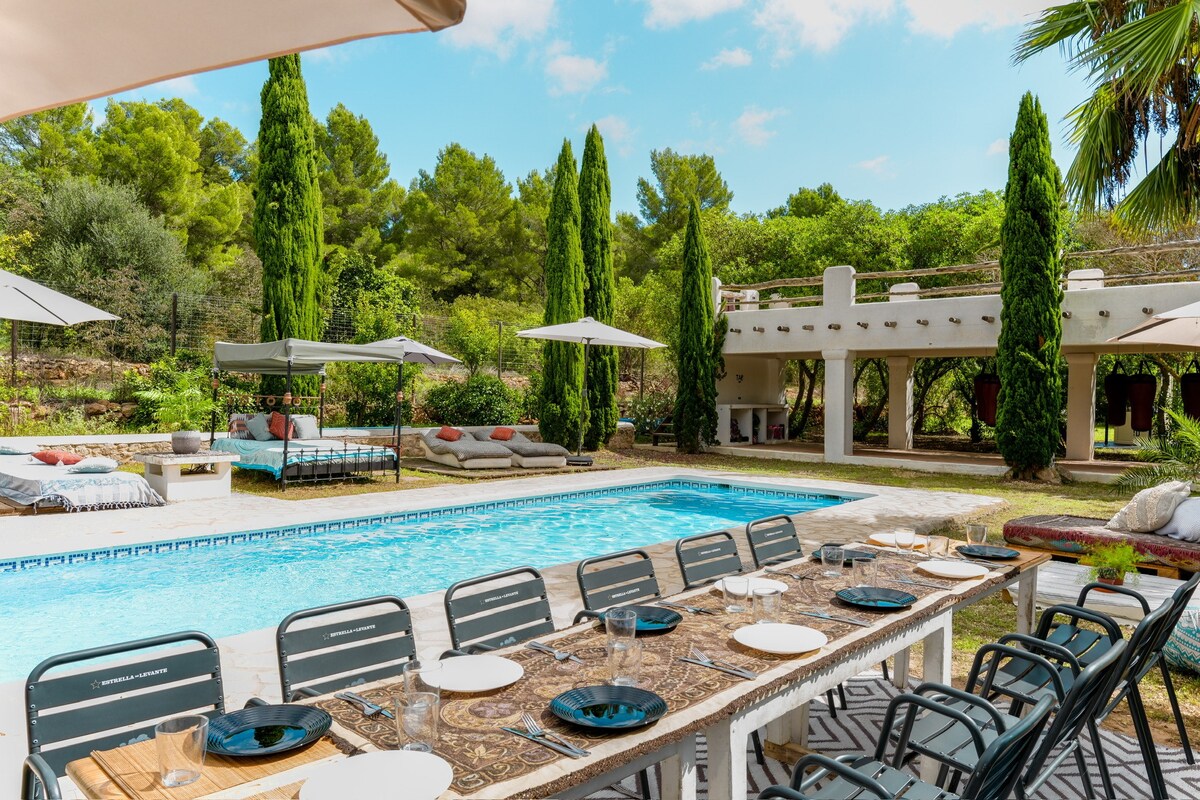 A spacious outdoor dining area with a long wooden table is set for a meal, surrounded by comfortable seating and shaded by large umbrellas. A sparkling swimming pool is visible, with relaxation beds and lush greenery providing a serene backdrop.