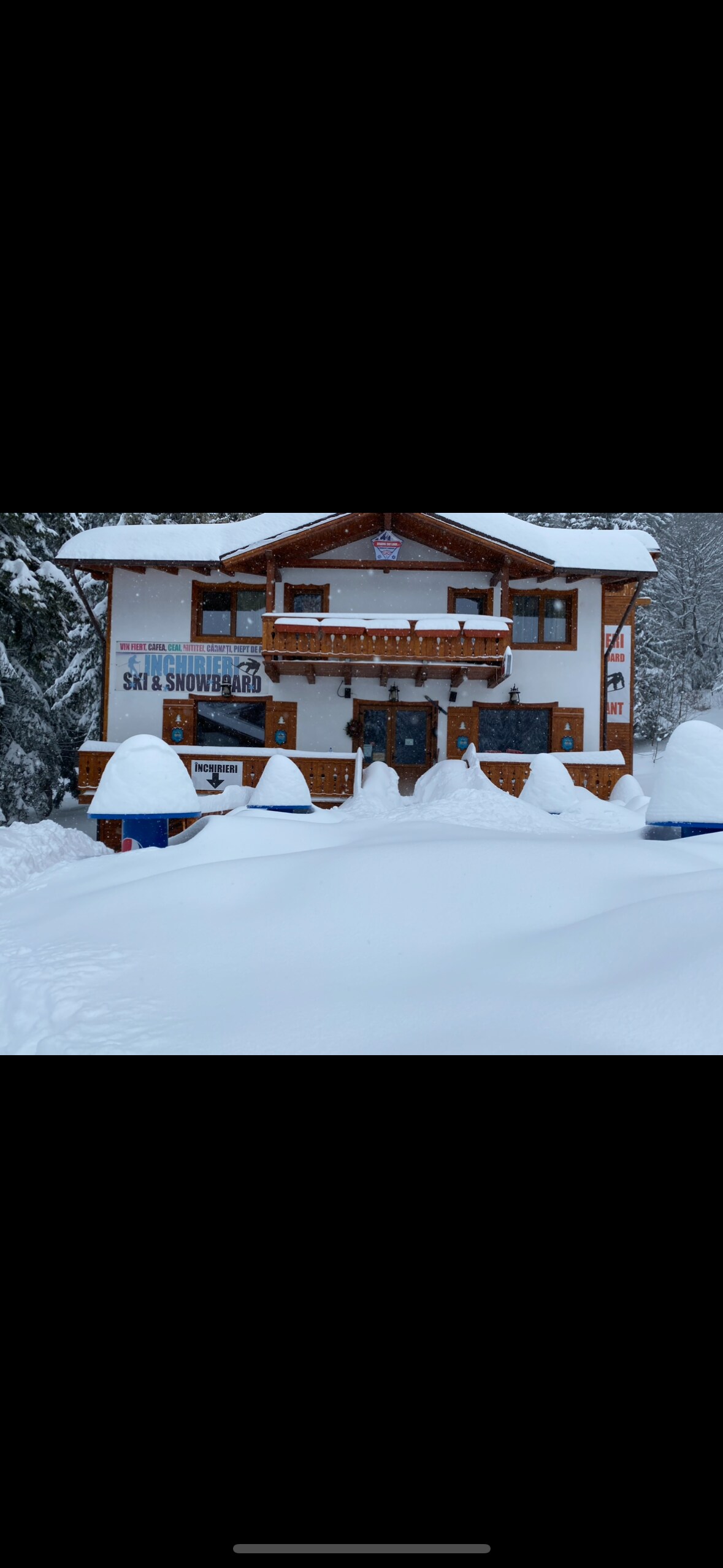 A charming wooden cabin is enveloped in a blanket of freshly fallen snow. The structure displays a welcoming facade with a large balcony and decorative elements. Snow-covered landscaping and seating areas are visible in the foreground, enhancing the wintry setting.