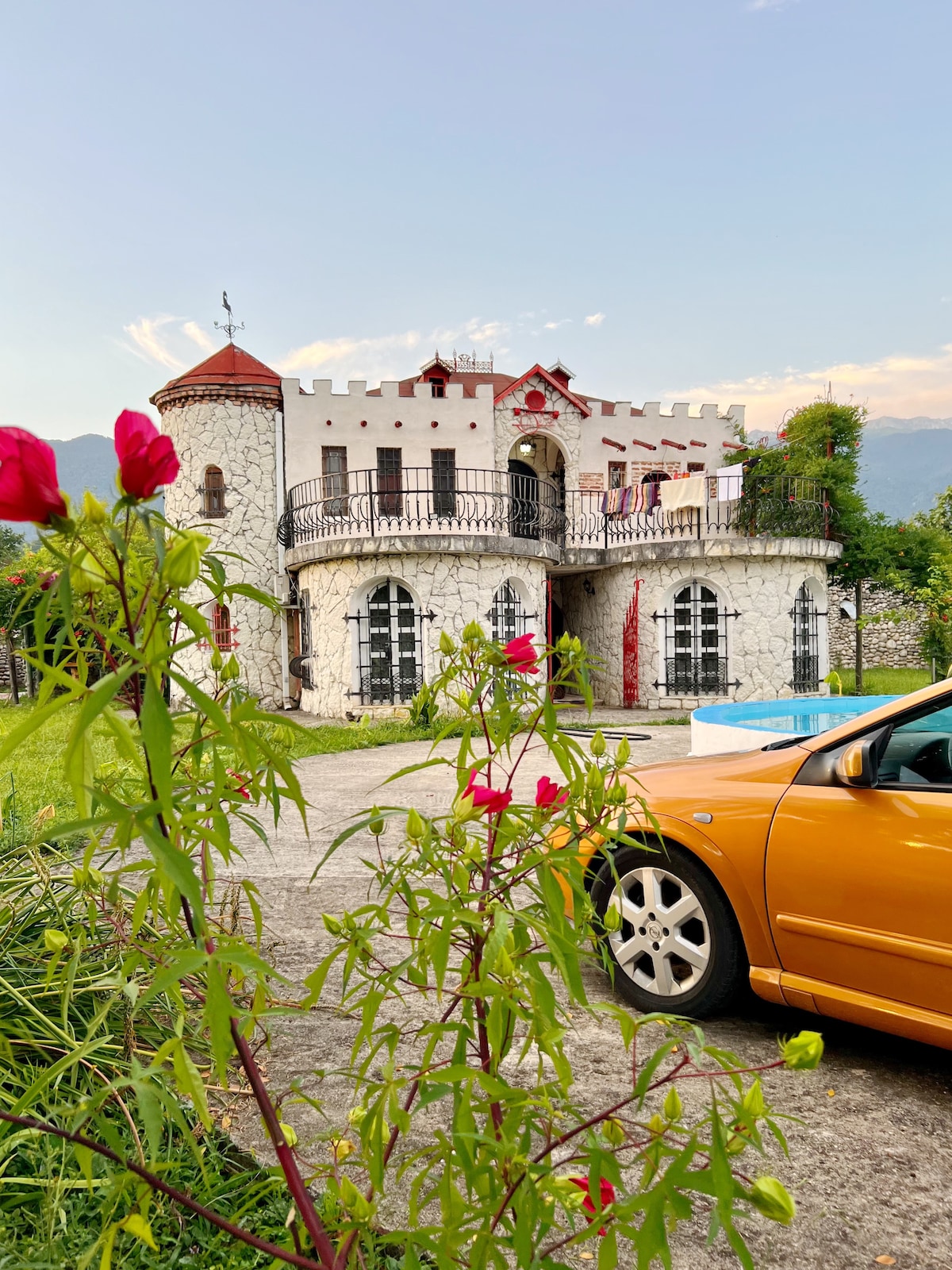 The villa, resembling a castle, is framed by vibrant flowers and green foliage. Its stone façade is accented by turrets and decorative arches, while a driveway leads to the entrance. The mountains serve as a serene backdrop under a clear sky.