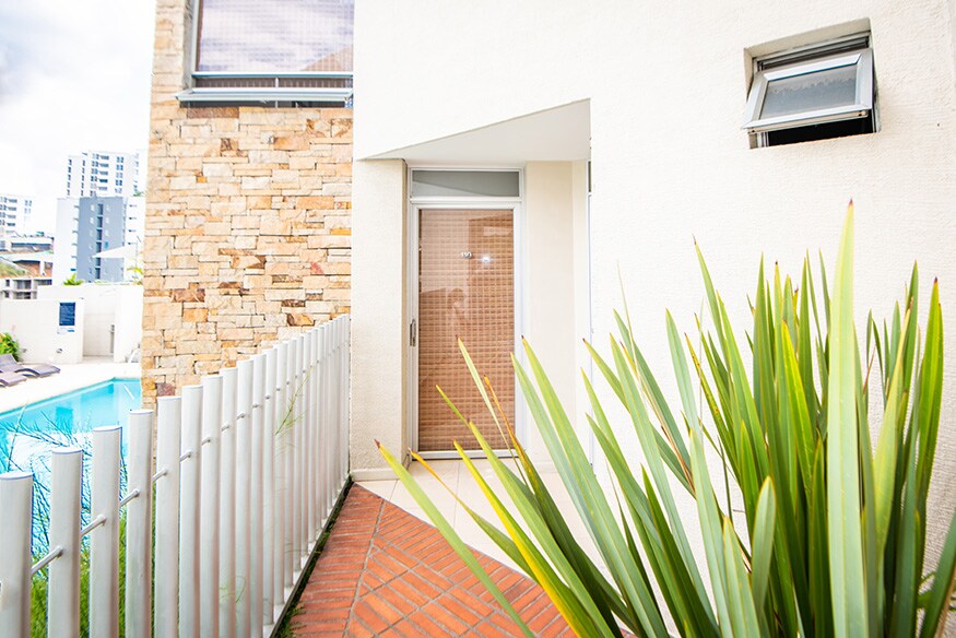 The exterior view showcases a modern entrance surrounded by greenery, with a light beige façade accentuated by a stone feature wall. A pathway leads to a door framed by a screen, while a swimming pool is visible in the background.