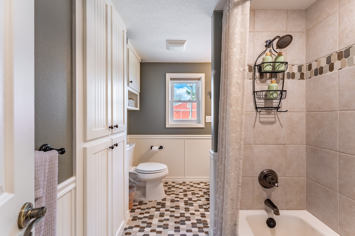 The bathroom features neutral tiled flooring with an attractive pattern. A tub is positioned next to a shower area containing a hanging caddy of toiletries. Cabinets provide storage space, complemented by a window that allows natural light, showcasing the view of the outdoors.