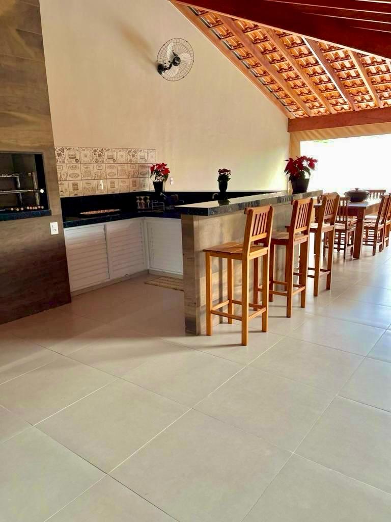A modern kitchen area features a spacious countertop with bar stools arranged around it. The high ceiling showcases a wooden beam structure, while bright natural light floods the space through large windows. Decorative plants are placed atop the countertop, enhancing the inviting environment.