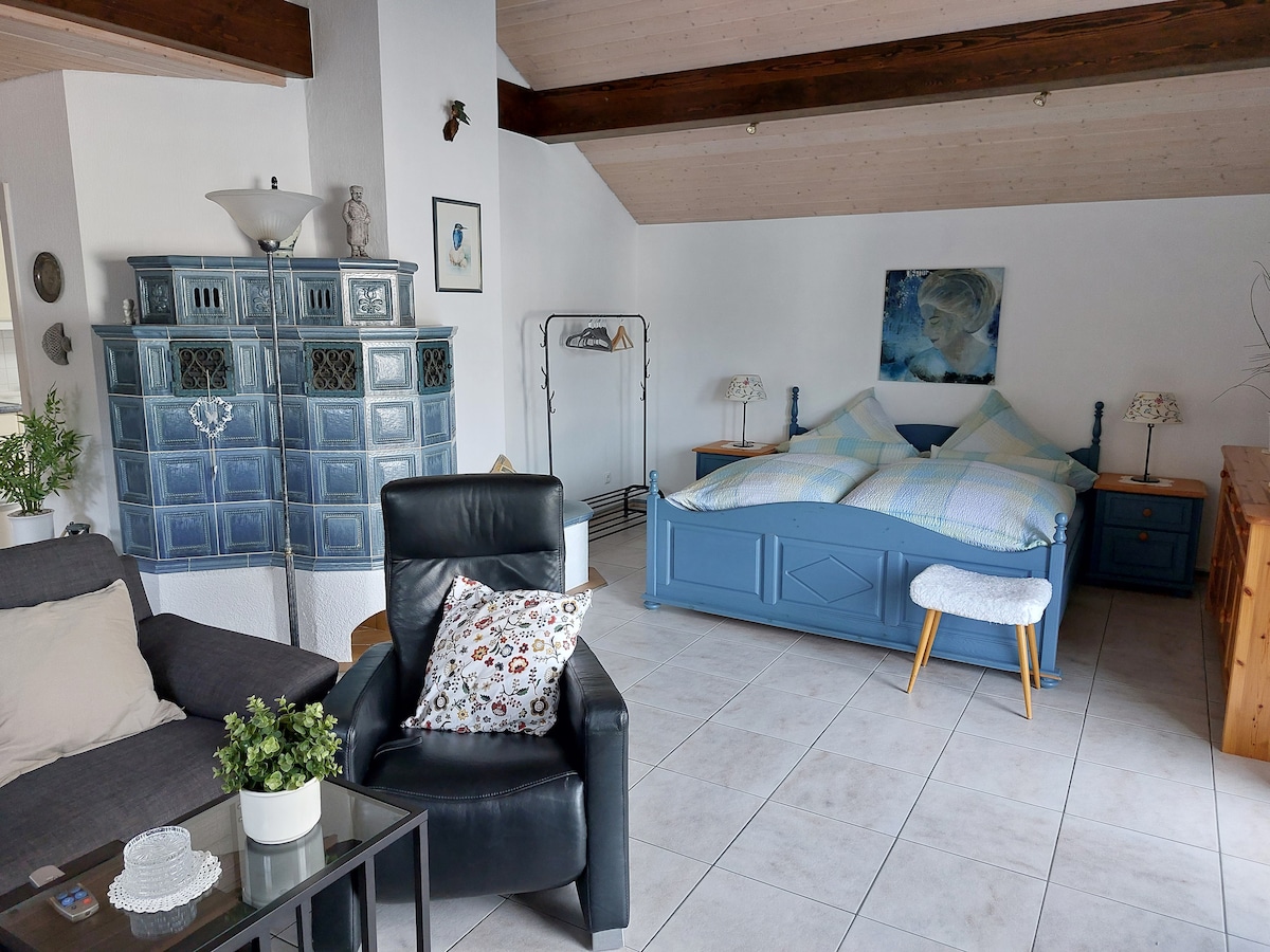 A spacious living and sleeping area is featured, with a cozy double bed adorned with soft bedding. A comfortable armchair and a small table provide a relaxing space, while a classic blue cabinet adds character to the room. Light is softly filtered through the windows.