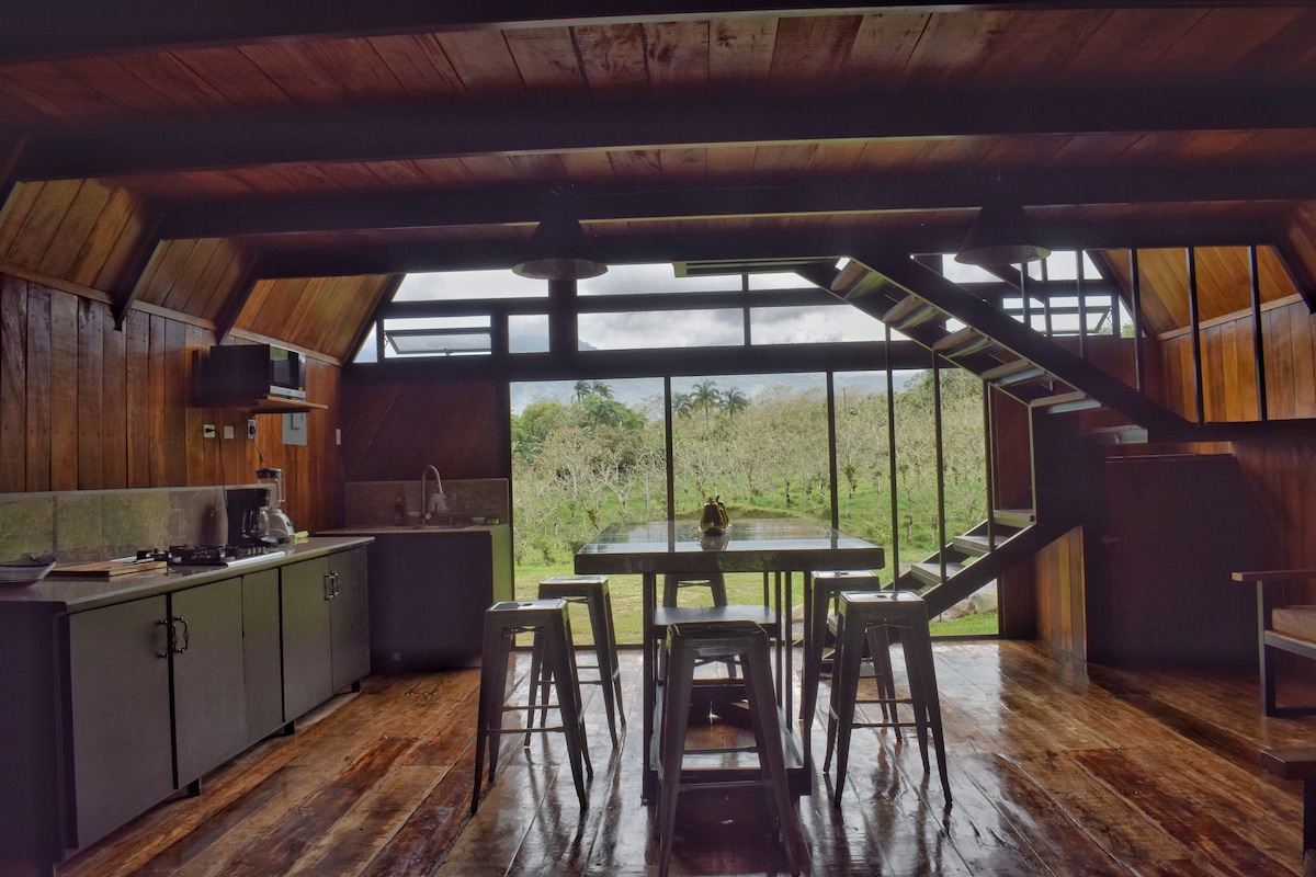An open-concept kitchen and dining area features wooden walls and spacious hardwood floors. The kitchen is equipped with modern appliances. A large table with several stools is positioned centrally, while a staircase leads to the upper level. Natural light fills the space through expansive windows.