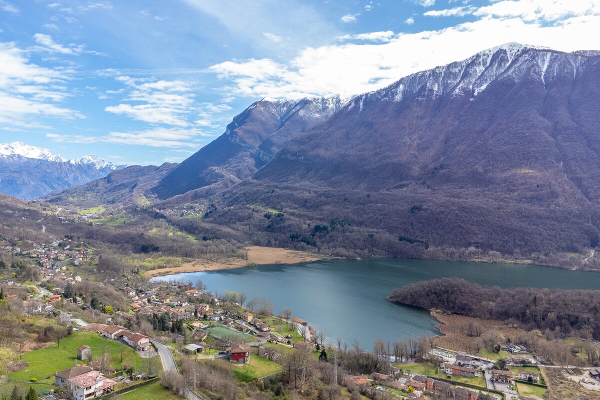A panoramic view captures the serene lake surrounded by mountains, with snow-capped peaks and green hills in the distance. The landscape features a mix of residential buildings and natural vegetation, highlighted by a clear sky above.