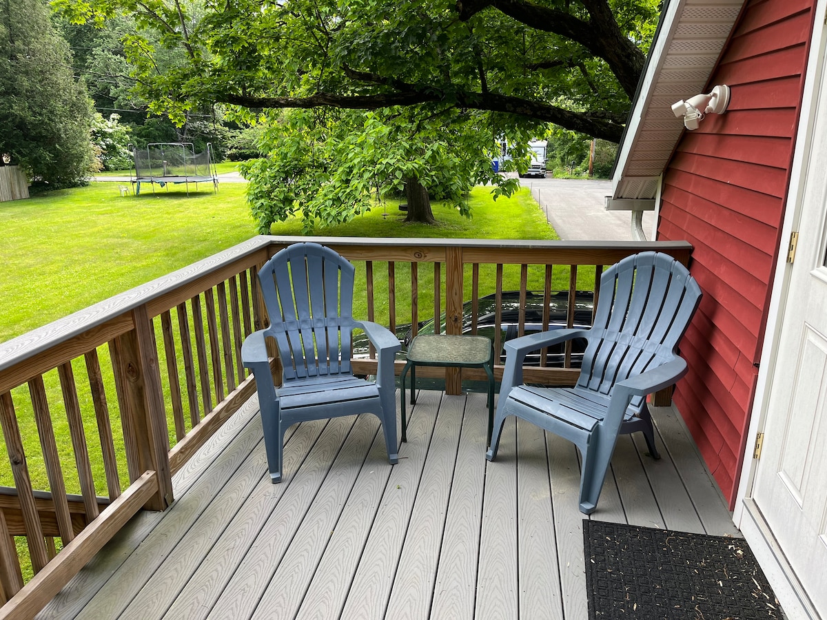 A private deck features two blue Adirondack chairs and a small glass table, positioned beside lush greenery and a large tree. The deck's wooden railing provides a view of the garden and surrounding landscape.