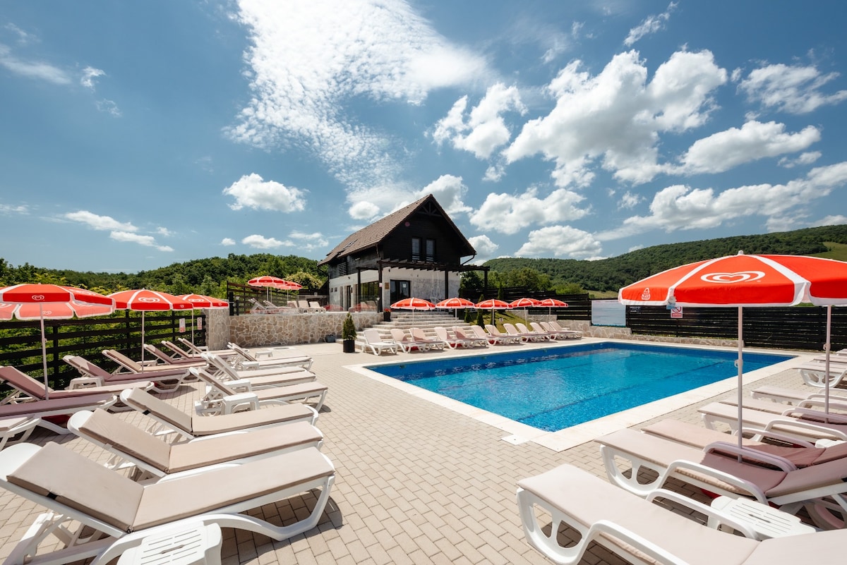 An outdoor swimming pool is surrounded by sun loungers, each paired with an orange umbrella. A two-story house with a thatched roof is situated in the background, framed by lush greenery and a blue sky filled with scattered clouds.