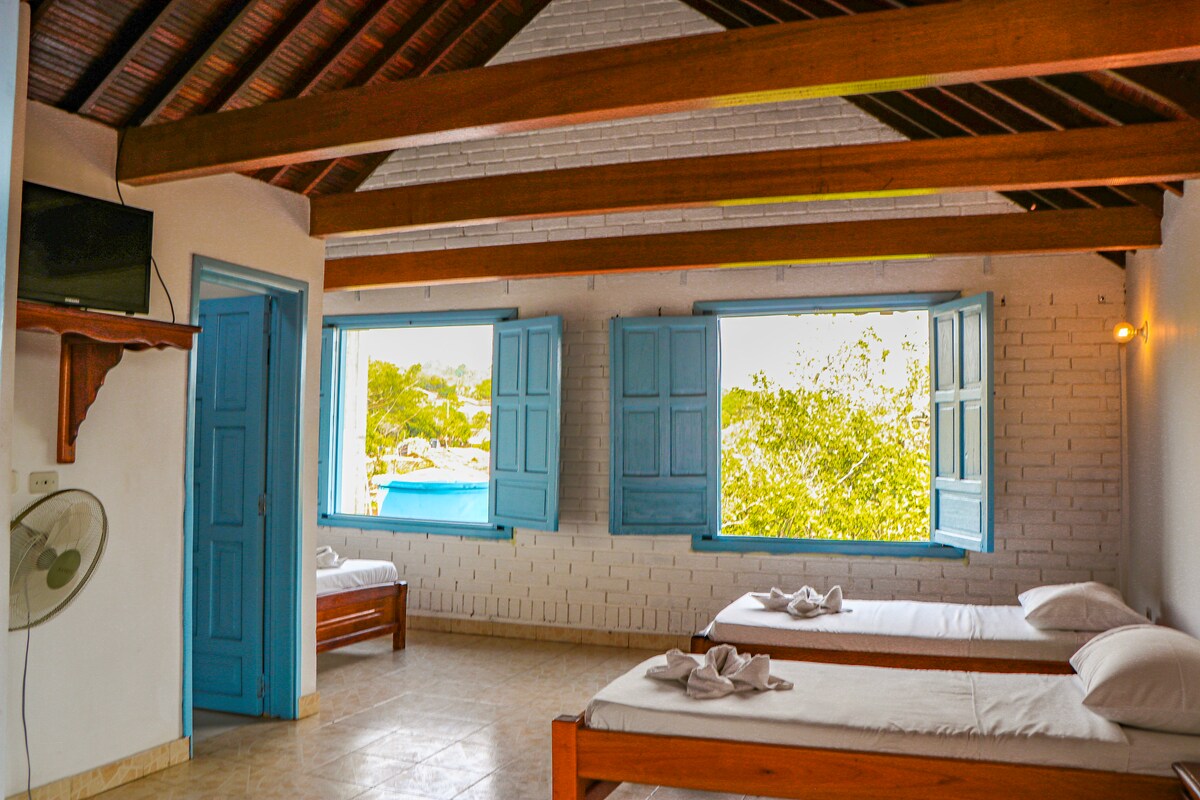 A bright sleeping area features two wooden beds adorned with neatly arranged towels. Blue windows are opened to allow natural light and fresh air, while a ceiling fan circulates the air. Light-colored walls and a wooden ceiling create a spacious atmosphere.