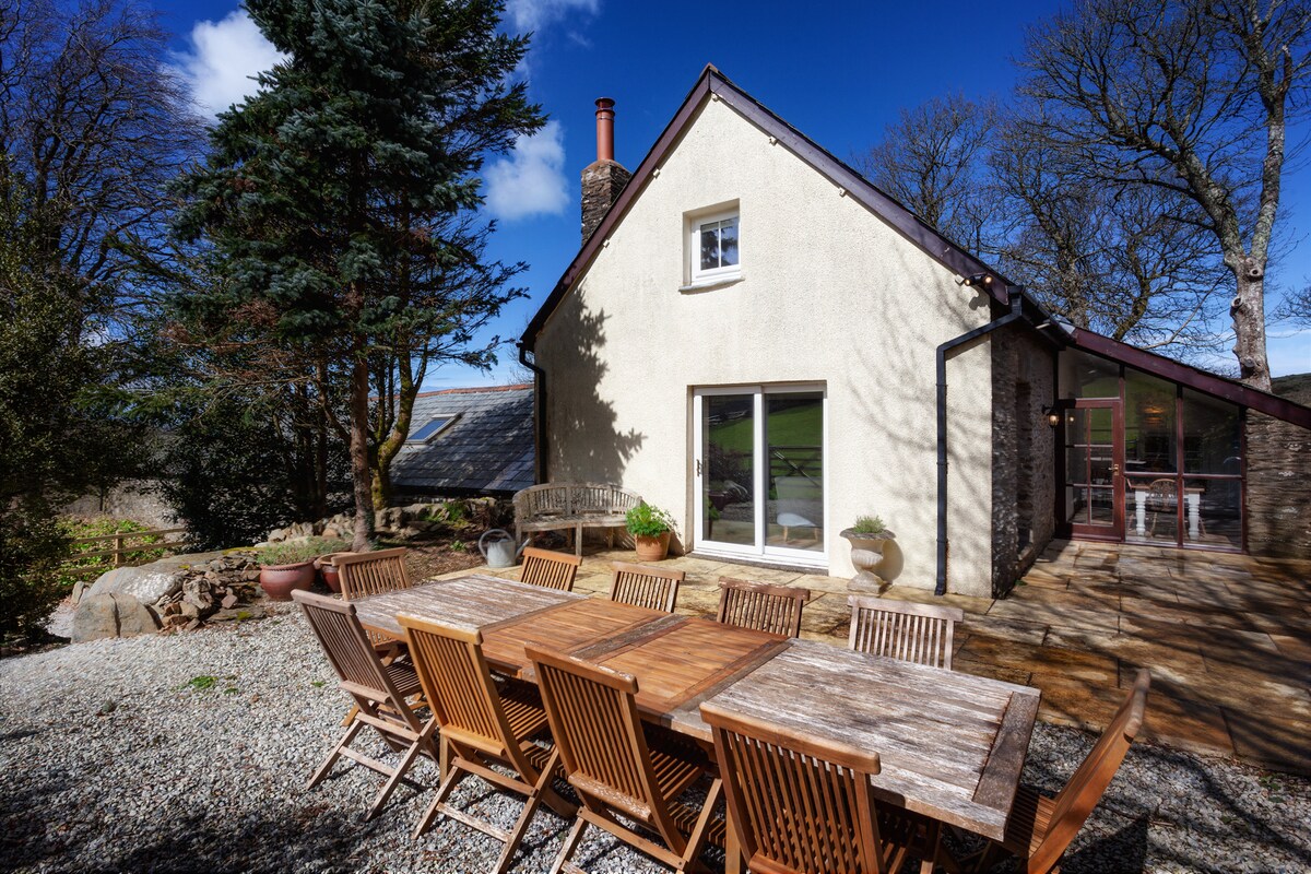 An outdoor dining area is displayed featuring a long wooden table surrounded by wooden chairs. The house is visible in the background, showcasing a simple exterior and large glass sliding doors that open onto the terrace. Lush trees frame the scene under a clear blue sky.