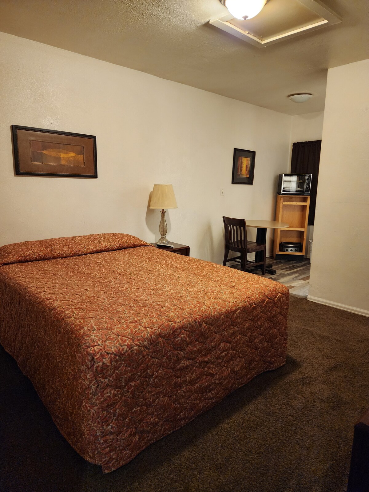 A cozy room features a spacious bed with a patterned quilt, complemented by soft lighting from a nearby lamp. A small wooden table and chair are positioned against the wall, with framed artwork adding subtle decor to the space.