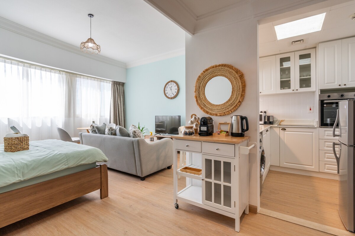 A bright studio space features a queen-sized bed with soft bedding next to a cozy seating area. A modern kitchen is visible, equipped with essential appliances. Large windows allow natural light to fill the room, enhancing its welcoming atmosphere.