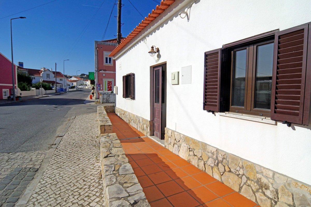 A charming exterior view shows a single-story cottage with a white facade and brown shutters. A stone walkway leads to the entrance, while the surrounding area features paved pathways and colorful buildings across the street under a clear blue sky.