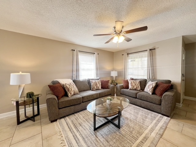 A spacious living area is furnished with a gray sectional sofa and a matching loveseat, both adorned with decorative pillows. A round glass coffee table sits in the center, complemented by a soft area rug. Light enters through two windows, enhancing the relaxed atmosphere.