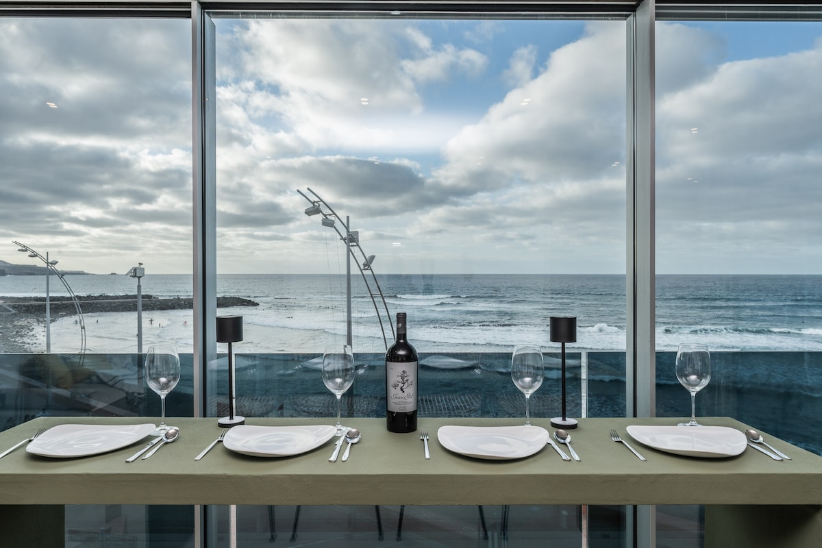 A dining table is set for four, featuring elegant plates and wine glasses. Beyond the glass railing, sweeping views of the beach and ocean are visible, with gentle waves lapping against the shore. A bottle of wine adds a touch of sophistication to the scene.