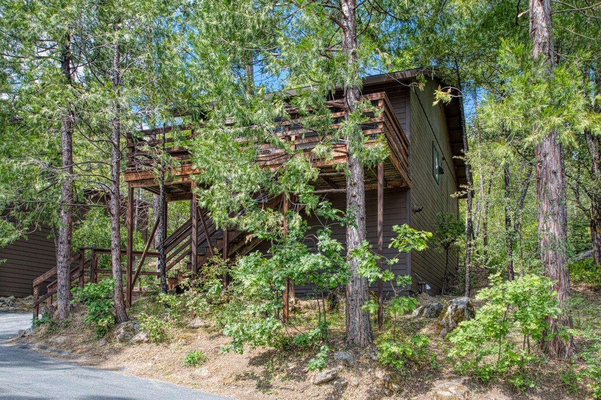 The two-story cabin is surrounded by tall pine trees, with a wooden deck visible on the upper level. Steps are leading to the entrance, which is partially obscured by greenery. The natural landscape enhances the cabin's secluded setting.