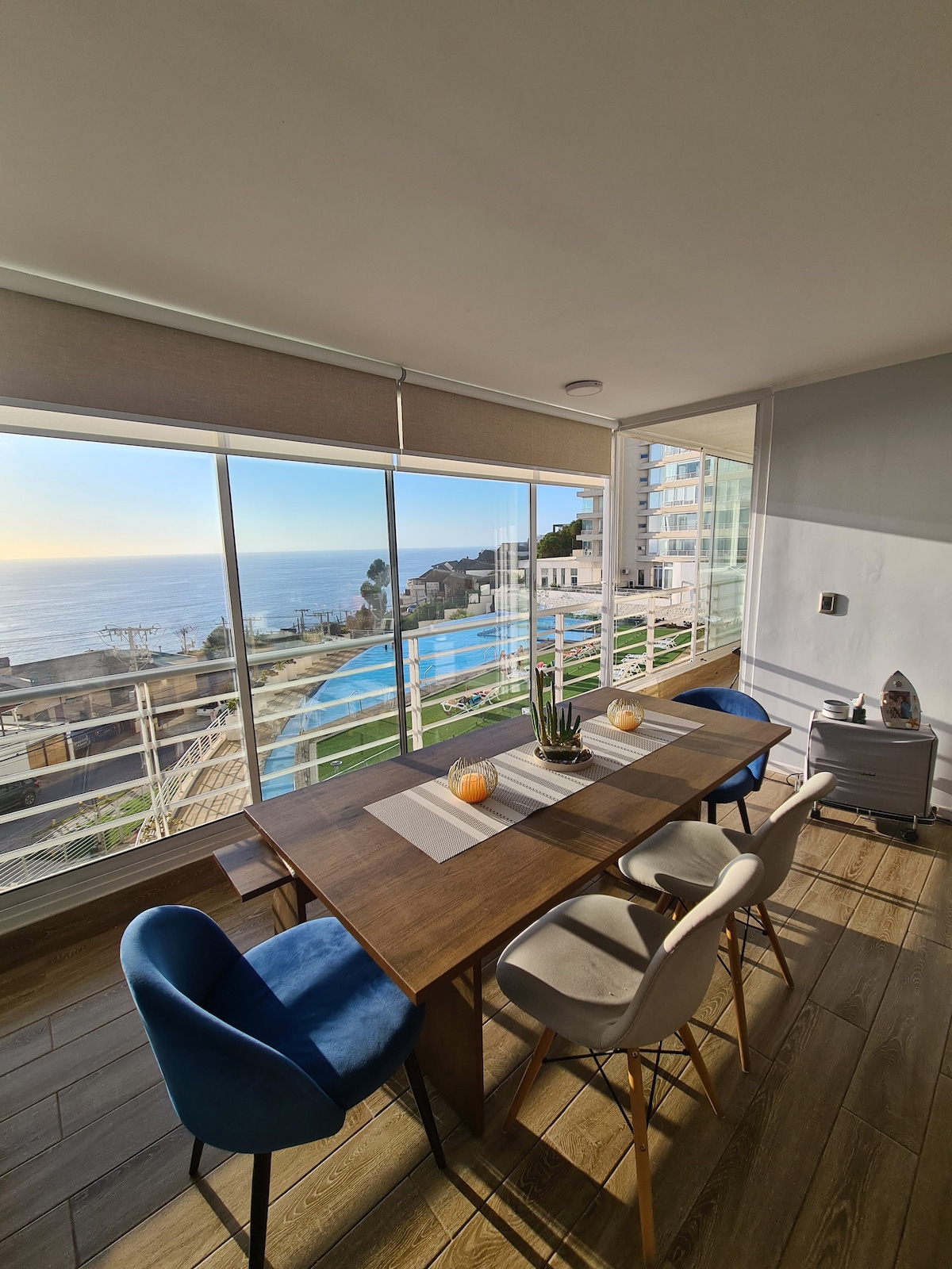 A dining area is illuminated by natural light, featuring a wooden table surrounded by four chairs in varied colors. A large window spans the wall, offering expansive views of the ocean and surrounding landscape, while soft fabric shades filter the sunlight.