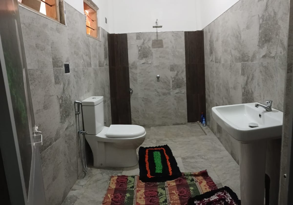 A spacious bathroom features natural stone walls and a mix of light and dark tiles. A modern toilet and sink are positioned alongside each other, while a colorful woven rug adds a touch of warmth to the tiled floor. Light enters through small windows.