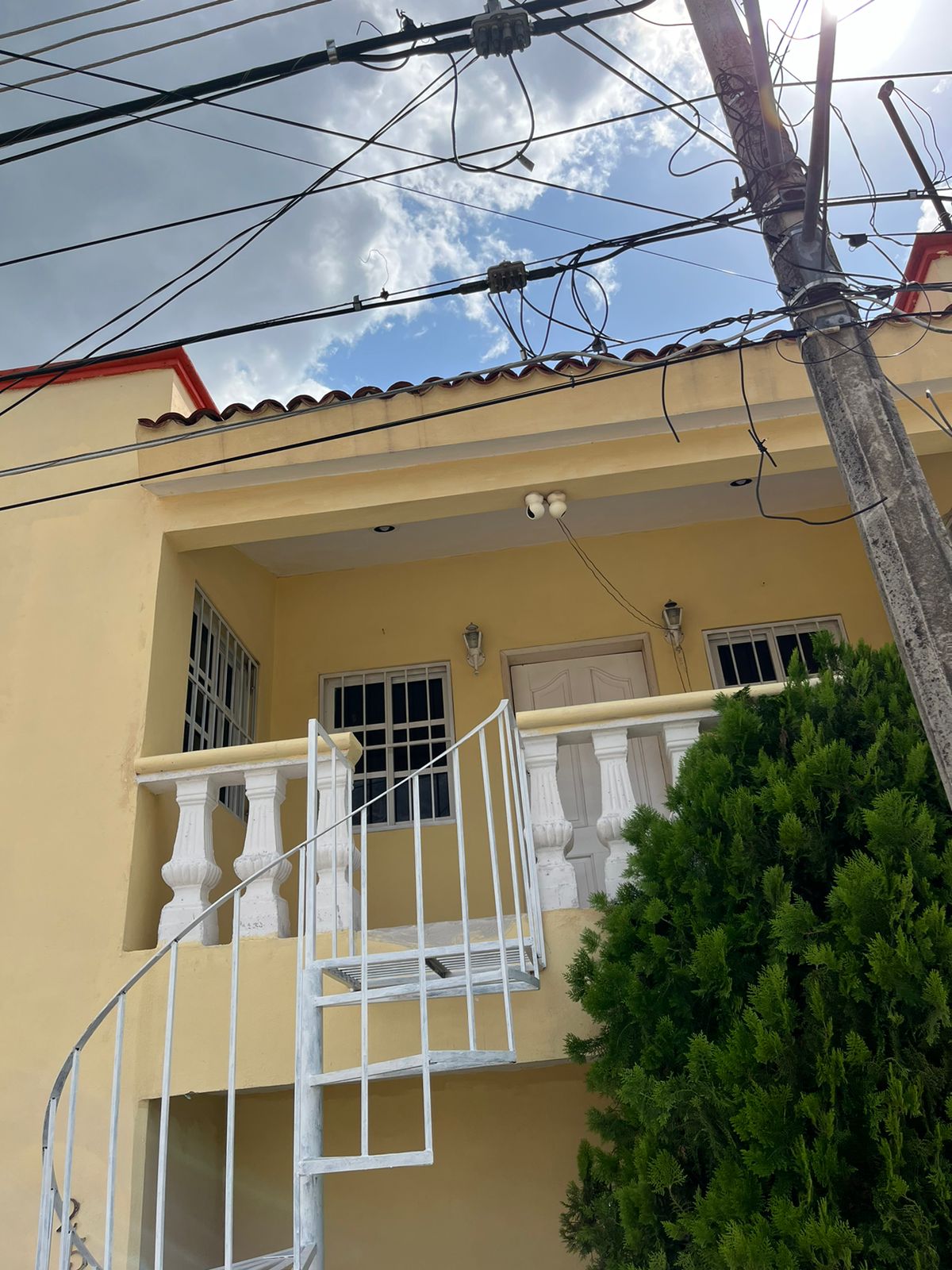 A two-story house is showcased with a yellow exterior and a red roof. A white spiral staircase leads to the upper level, while large windows allow natural light to fill the space. Nearby greenery softens the urban setting.
